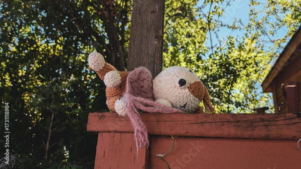 Fototapeta Close up of a cute crocheted stuffed animal abandoned on a fence with a tree and branches in the background. 