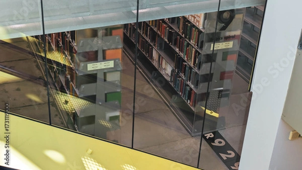 Fototapeta High angle view of nonfiction book stacks as seen through several panes of glass and partially hidden by a structural beam painted yellow.  