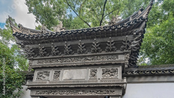 Fototapeta Ancient Chinese architecture. A fragment of a building with curved roof edges, carvings on cornices. Green branches of trees against a background of blue sky and clouds. China. Yu Garden. Yuyuan