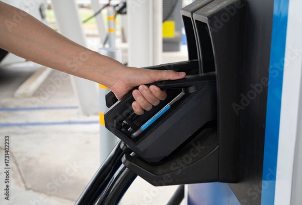 Fototapeta Hand holding electric plug connector at public EV charging station for sustainable vehicle battery power. Modern green technology infrastructure supporting clean energy transportation solutions.