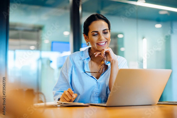 Obraz Smiling woman in office using laptop while holding glasses and working thoughtfully