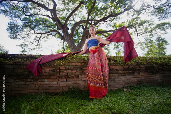 Fototapeta Woman in traditional attire poses gracefully with red scarf in front of ancient brick wall and large tree