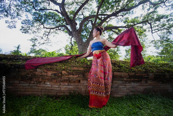 Fototapeta Traditional Thai dancer in colorful attire poses gracefully by ancient brick wall under large tree