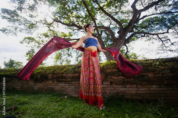 Fototapeta Traditional Thai dancer in colorful attire gracefully poses with flowing fabric in lush garden setting