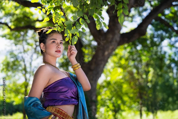 Fototapeta Woman in traditional attire stands gracefully under tree, surrounded by lush greenery and sunlight