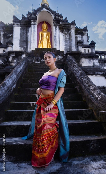 Fototapeta Traditional attire woman stands on ancient temple steps, golden Buddha statue in background. Cultural heritage and serene atmosphere