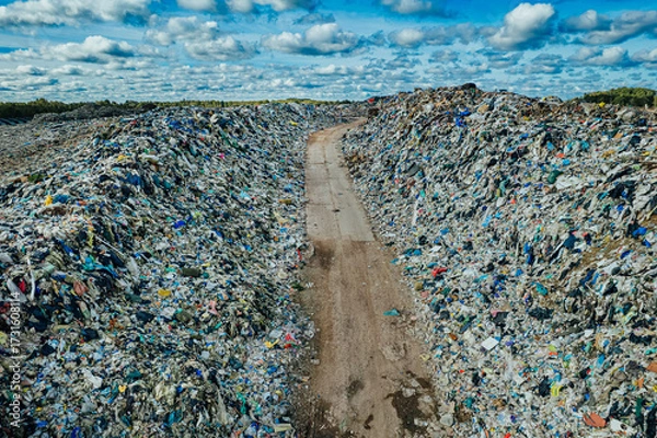 Fototapeta A dirt road cuts between massive piles of garbage in a landfill, symbolizing pollution, overconsumption, and the urgent global challenge of waste management
