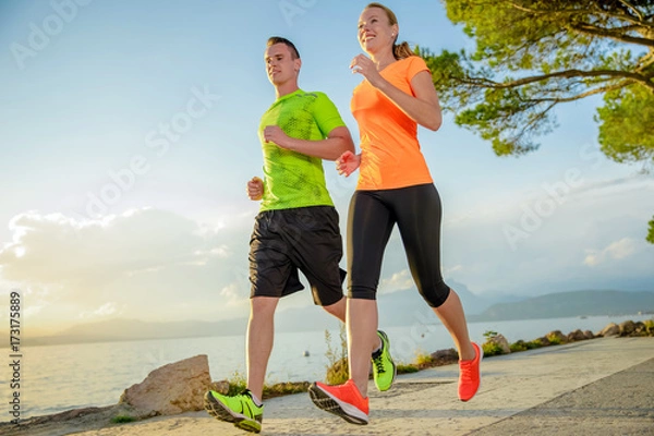 Obraz Young sexy couple is running along the promenade. they are doing their sport workout in the beautiful sundown along the beach. colorful dress, trees, water, mountains and a amazing blue sky.