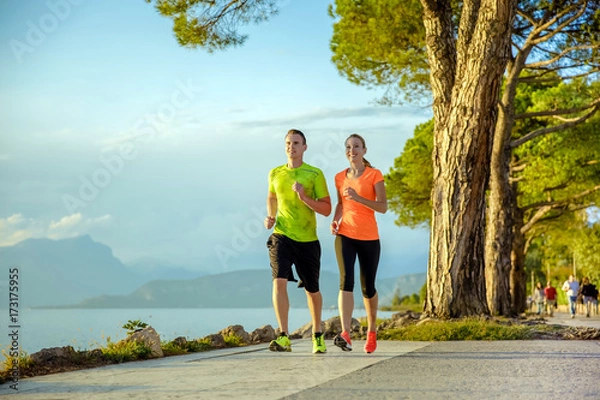 Obraz Young sexy couple is running along the promenade. they are doing their sport workout in the beautiful sundown along the beach. colorful dress, trees, water, mountains and a amazing blue sky.