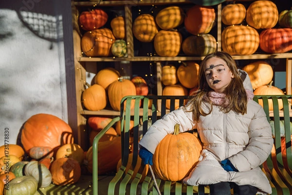 Fototapeta A young girl with a painted face sitting on a green bench, holding a pumpkin in front of a rustic wooden stand filled with pumpkins. Halloween concept