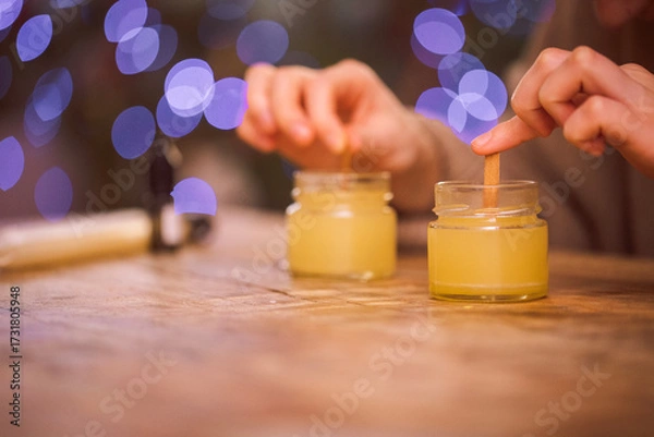 Fototapeta Hands of a person carefully stirring wax in glass jars on a rustic wooden table, with soft bokeh lights in the background, creating a cozy crafting atmosphere