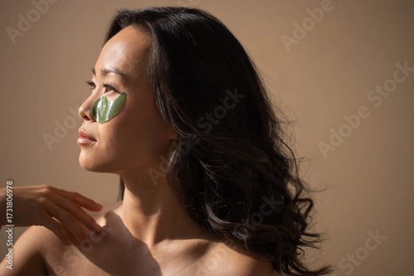 Fototapeta Asian woman with green facial mask, enjoying skincare routine, illuminated by soft natural light, showcasing beauty and self-care in a serene environment
