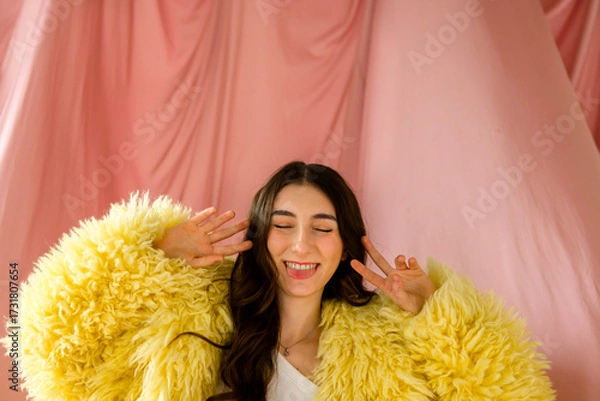 Fototapeta Young woman with long dark hair wearing a fluffy yellow coat is playfully posing against a soft pink backdrop, showcasing a joyful and vibrant atmosphere of self-expression
