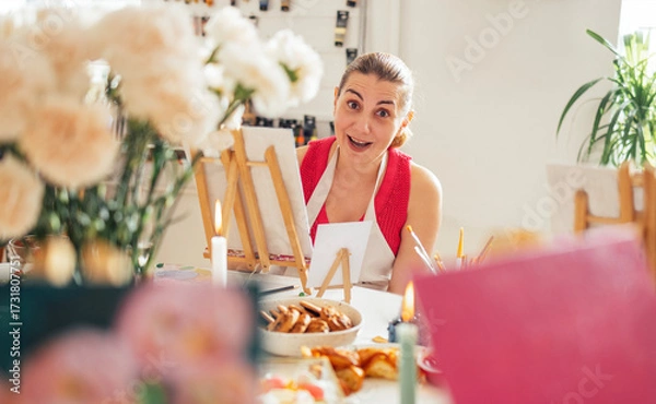 Fototapeta Female artist with brown hair, wearing an apron, is smiling at the camera in a bright studio filled with flowers, easels, and art supplies, showcasing a creative atmosphere