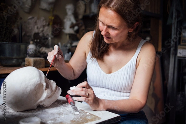 Fototapeta Female artist carefully painting a sculpted head in a creative studio, surrounded by artistic tools and materials, showcasing dedication to the craft of sculpture and artistry