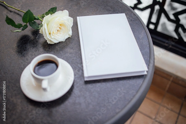 Fototapeta White rose placed beside a blank notebook and a cup of coffee on a round table, creating a serene atmosphere for relaxation and reflection in a cozy setting