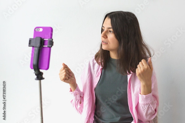 Fototapeta Woman in pink shirt speaking to smartphone on tripod, gesturing with hands and looking engaged, possibly recording vlog or video message, concept of communication