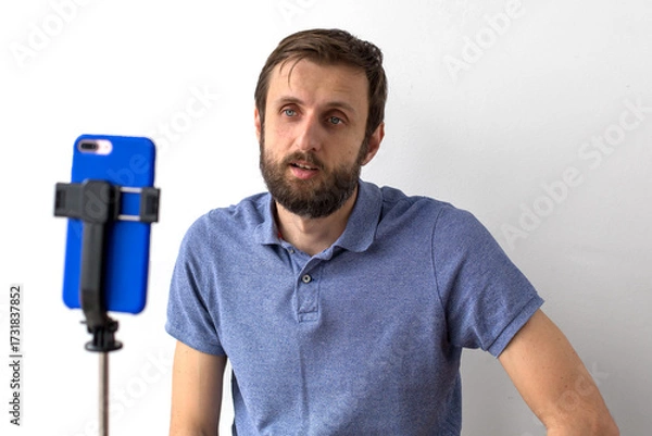 Fototapeta Man with beard in blue polo shirt sitting in front of smartphone on tripod, recording video against white wall, concept of communication