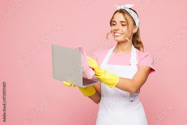 Fototapeta Young IT woman she wear white apron hold basin with detergent bottles while doing housework tidy up wiping laptop pc computer screen with cloth isolated on plain pink background. Housekeeping concept.