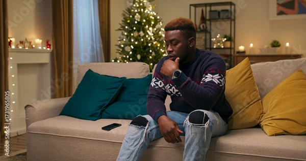 Obraz Sitting on a sofa, a young student of multi-racial background receives news that his flight home was canceled. He tosses the phone aside and bows his head in frustration.
