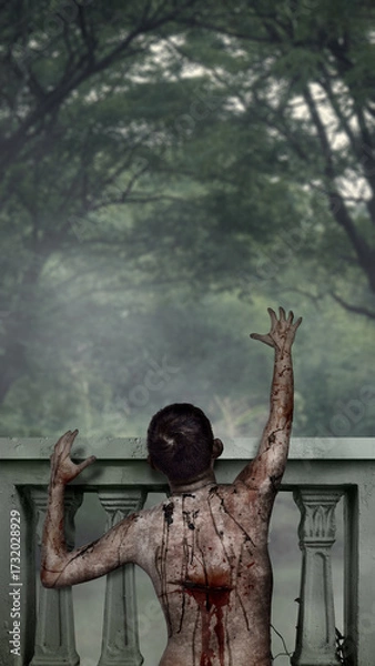 Fototapeta Scary male zombie with a wound and blood reaching out from the balcony railing for haunting. The scene is scary and creepy. Dramatic smoke or fog effect. Concept of Spooky Halloween