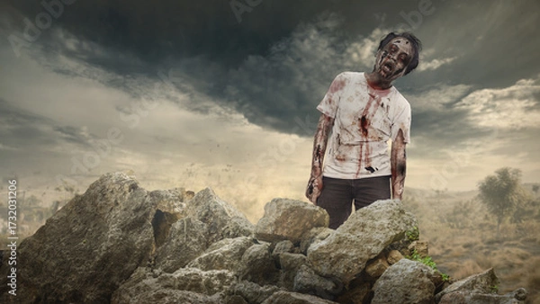 Fototapeta Scary male zombie with a wound and bloody walking on rocky terrain. The scene is scary and creepy. Dramatic sky. Concept of Spooky Halloween