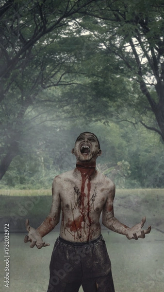 Fototapeta Scary male zombie with a wound and blood walking on the street surrounded by trees. The scene is scary and creepy. Dramatic smoke or fog effect. Concept of Spooky Halloween