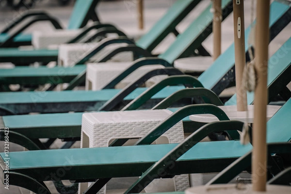Fototapeta Row of empty turquoise lounge chairs with white side tables at a resort poolside area, arranged neatly and ready for guests under sun umbrellas.