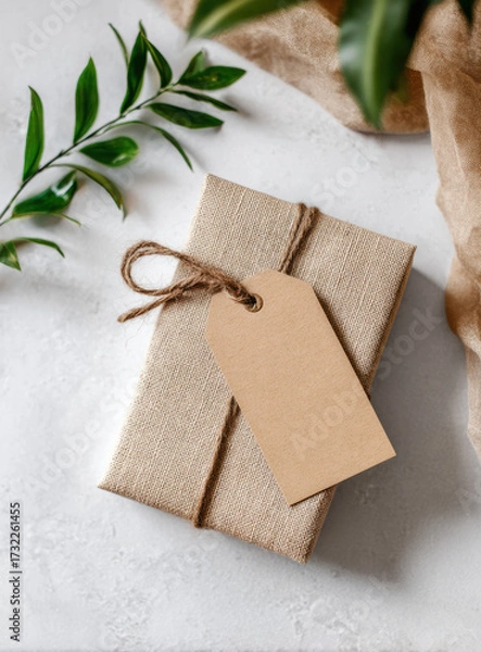 Fototapeta Thoughtful natural gift box present wrapped in rustic brown paper with blank tag. gentle composition with green leaf detail on white background for warm celebration
