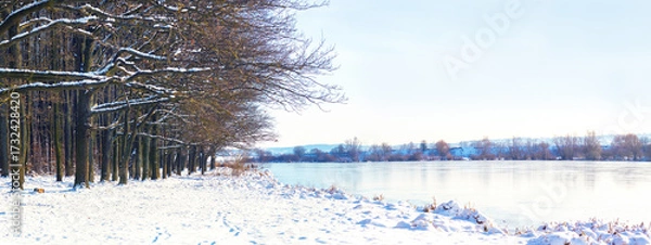 Fototapeta Panoramic winter landscape with a frozen lake, snow-covered banks, and trees illuminated by sunlight, creating a calm and majestic mood
