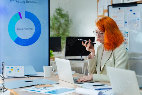Fototapeta Female data analyst using a smartphone to speak with an AI assistant or record voice note in a modern office.data meeting environment,ai assistant,futuristic tech,ai agent and future of AI technology