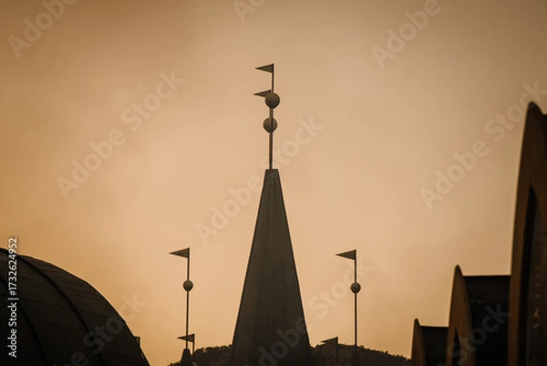 Fototapeta Silhouettes of pointed architectural structures with flags and spherical ornaments stand against a dramatic orange sunset sky, creating a moody and artistic view.