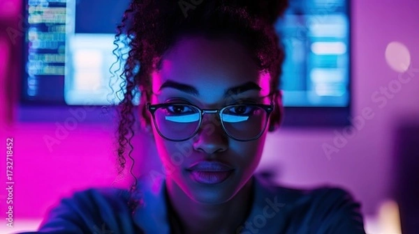 Fototapeta Focused young woman in glasses working at computer with neon purple and blue lighting screen code reflection in lenses