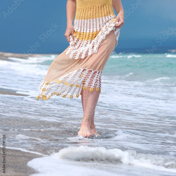Fototapeta Unrecognizable woman in a beautiful summer dress stands on wet sand near the water on the beach. Vacation at a seaside resort in the summer. Walking along the beach at sea, legs close-up