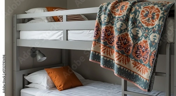 Fototapeta A grey bunk bed with white linens, orange and patterned pillows, a patterned blanket, and a wall-mounted lamp.