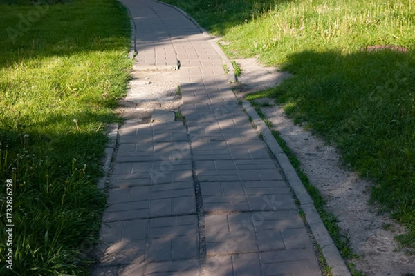Fototapeta Broken tiled path leading into the distance with grass on both sides, parallel dirt trail on the right, deep shadows and a bright sunlit patch across the path.