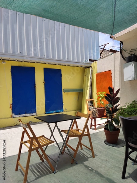 Fototapeta A vibrant and eclectic outdoor patio scene featuring a small black metal bistro table with two light wooden folding chairs, set against a bold, colorful architectural backdrop 