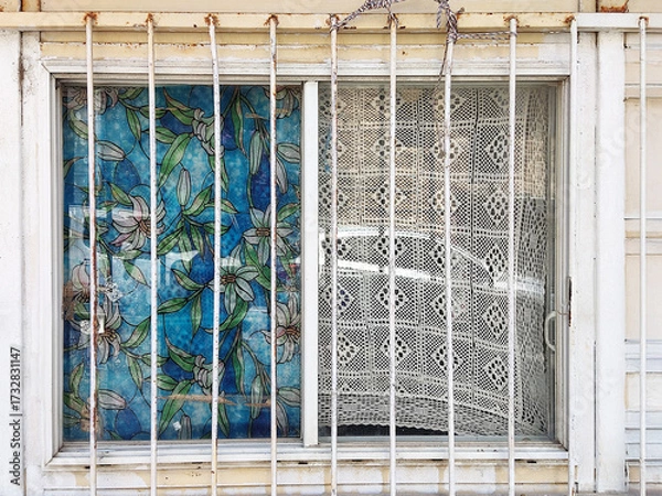 Fototapeta the left with vibrant floral stained glass, the right with a sheer crochet curtain. Rusty metal bars and a weathered wall frame the window, blending bright patterns with aged, neglected textures.