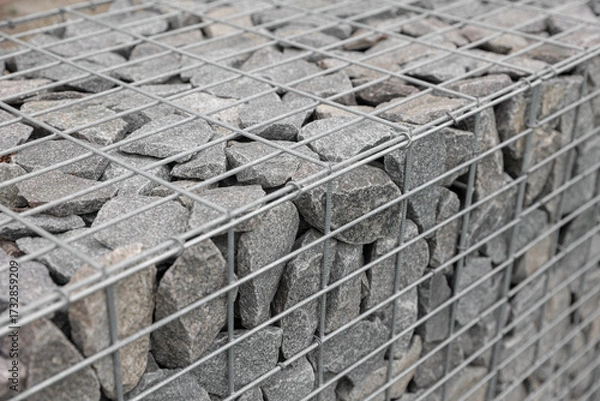 Fototapeta Stone fence in steel mesh and green lawn. Close up of gabion with tightly packed stones of varying sizes and textures. Combination of natural materials and industrial design.