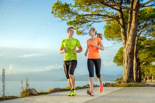Obraz Young sexy couple is running along the promenade. they are doing their sport workout in the beautiful sundown along the beach. colorful dress, trees, water, mountains and a amazing blue sky.