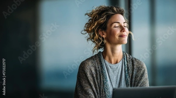 Fototapeta A woman with curly hair wearing a gray sweater, sitting in front of a laptop with her eyes closed, appearing to be meditating or deep in thought.