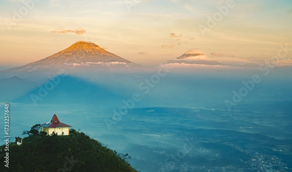 Fototapeta A small building sits atop a hill overlooking a valley with distant mountains shrouded in mist during a warm, golden sunrise.