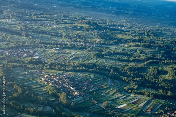 Fototapeta Aerial view of lush green rice terraces and a small village nestled in a valley, bathed in soft morning light.