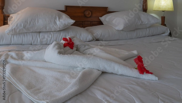 Fototapeta Romantic bed preparation for bedtime. On the white sheets, towels are folded in the shape of a heart and decorated with bright red hibiscus flowers. Pillows at the headboard. The bedside lamp is lit.