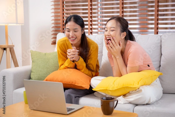 Fototapeta Two young asian woman sitting on sofa using laptop computer for video call in living room at home, female using social media on notebook for online and conversation distant, lifestyles concept.