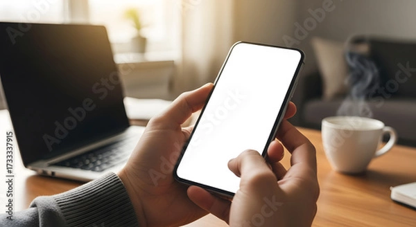 Fototapeta Hands holding a modern smartphone with a blank white screen in a well-lit room, with a laptop and a steaming cup of coffee on the table