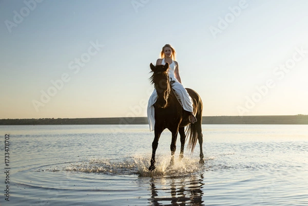 Obraz A woman in a flowing white dress rides her horse through the shimmering waters of Kuyalnik Lake, bathed in the golden evening light.