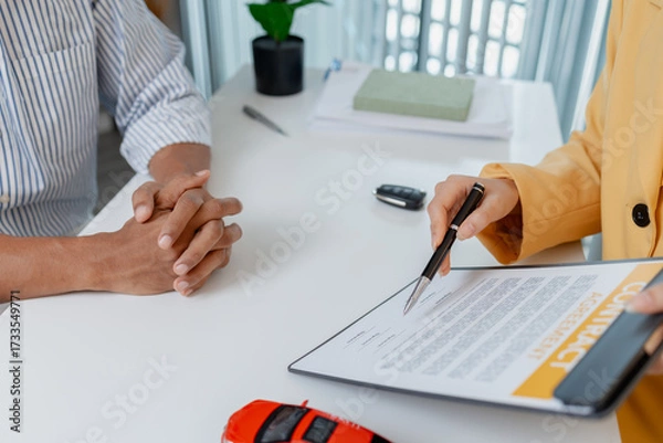 Fototapeta A car sales negotiation takes place in an office where a female salesperson in a yellow blazer explains details of a contract to a male client, showing car models and handing over keys.