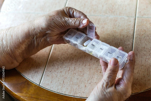 Fototapeta Elderly hands holding a weekly pill organizer with tablets and capsules, symbolizing health care, routine medication, treatment management, and daily wellness support