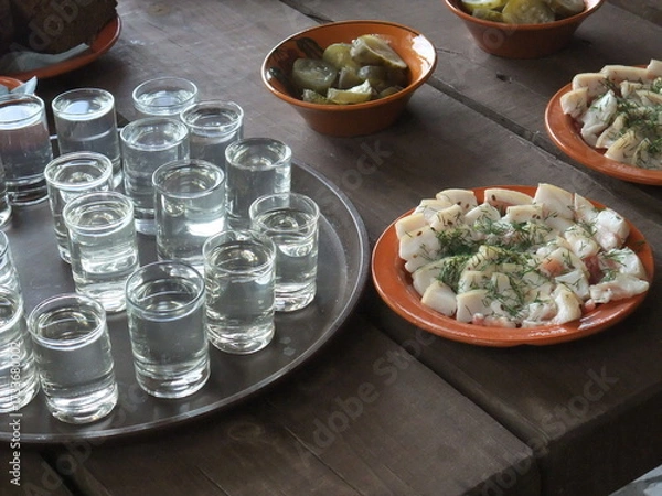Fototapeta Traditional Slavic table setting with vodka shots, salted lard with herbs, and pickles served on rustic wooden surface